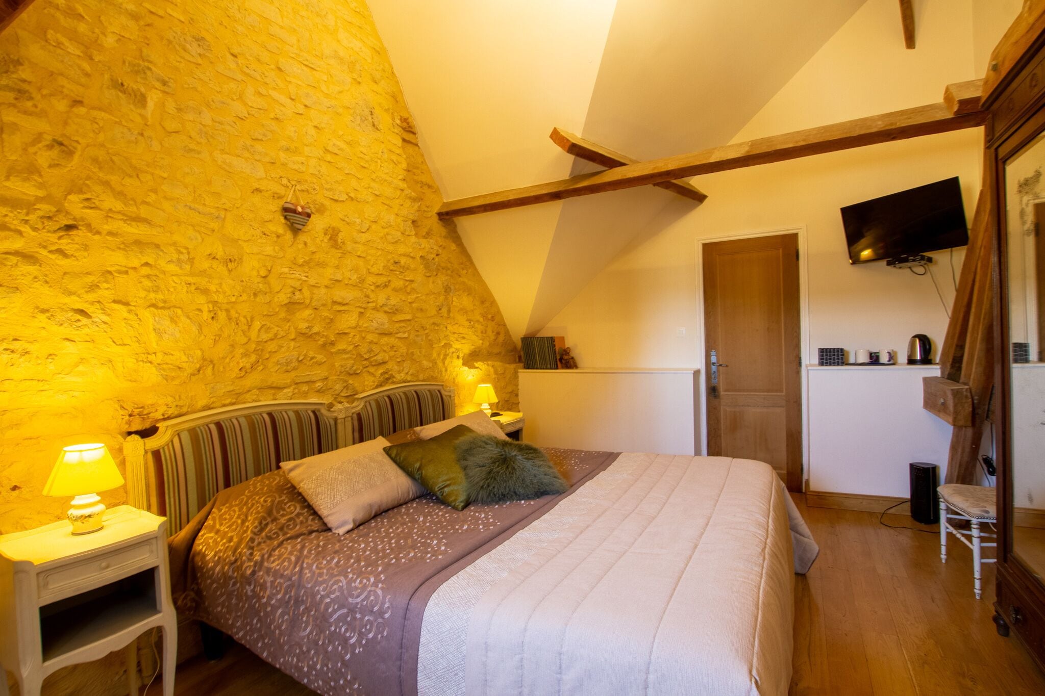 A bedroom with a stone wall, a bed with patterned bedding, a nightstand with a lamp, and a television mounted on the wall.