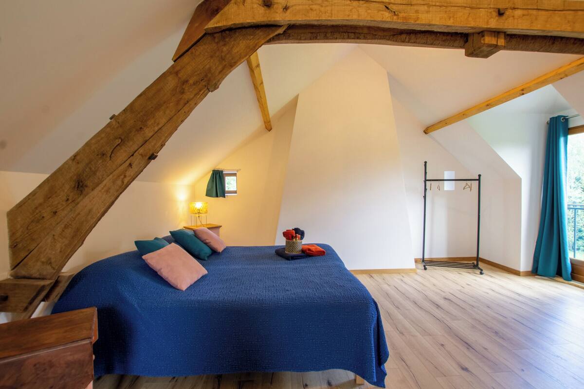 A bedroom with a sloped ceiling, a bed with a blue bedspread, pillows, a bedside table with a lamp, and a window with curtains.