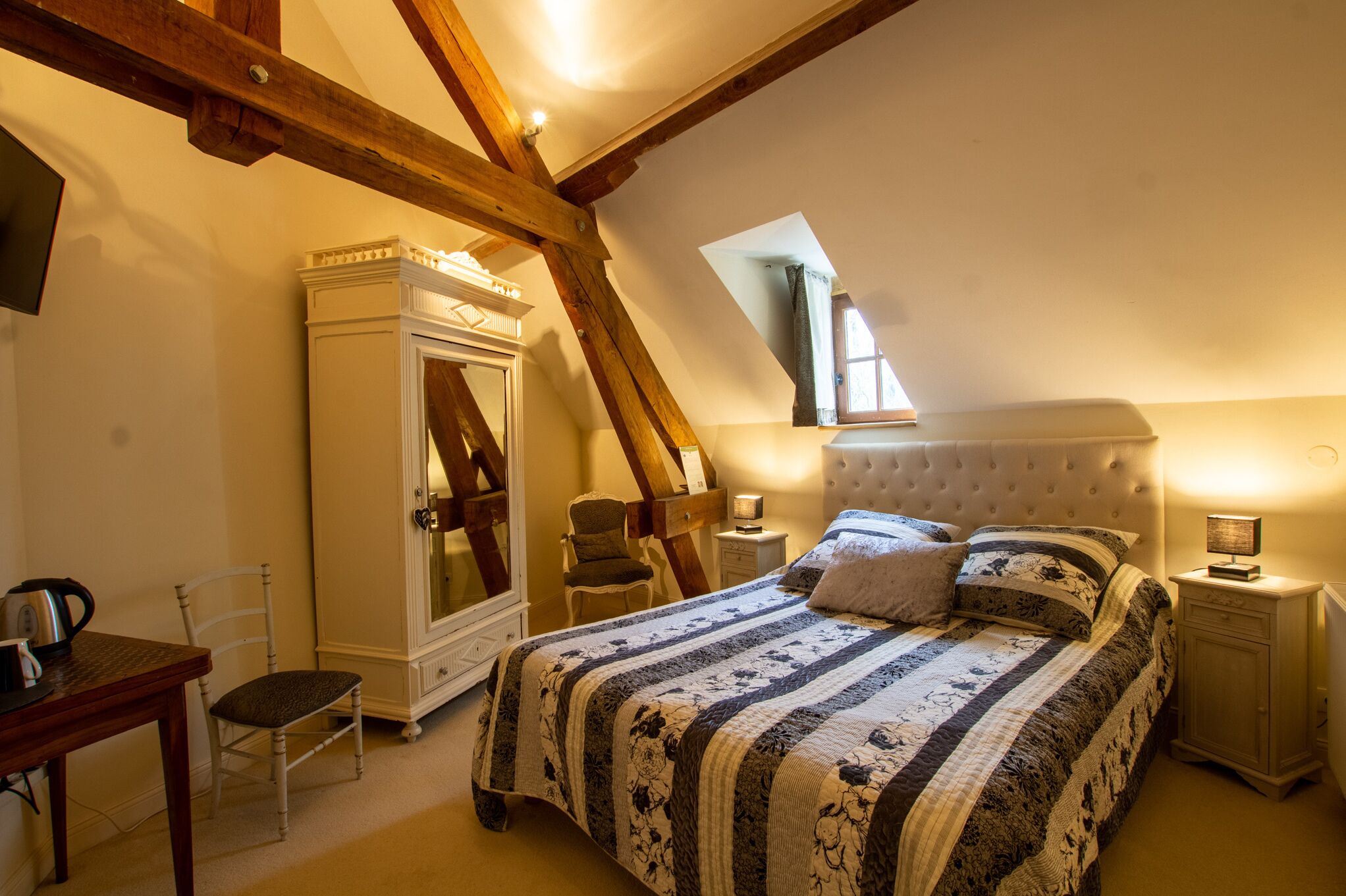 Un dormitorio en el ático con techo alto, vigas de madera a la vista, una cama grande con colcha floral, un aparador, un escritorio y una silla.