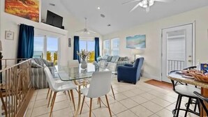 A spacious living area with a glass dining table, white chairs, a blue sofa, and a ceiling fan.