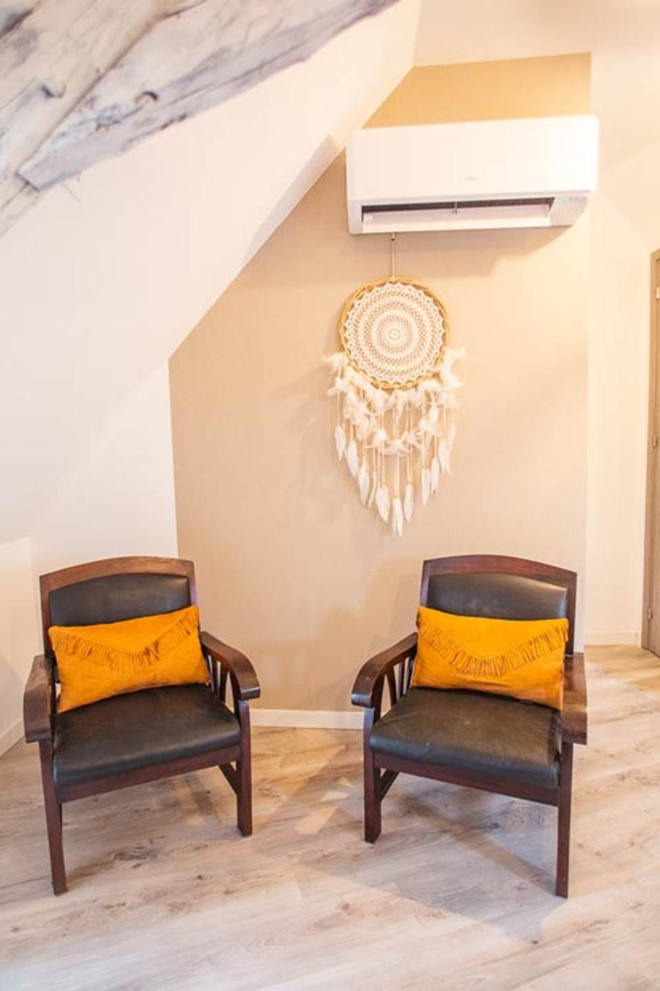 Two wooden chairs with yellow cushions, a wall-mounted dreamcatcher, and an air conditioning unit in a room with a sloped ceiling.