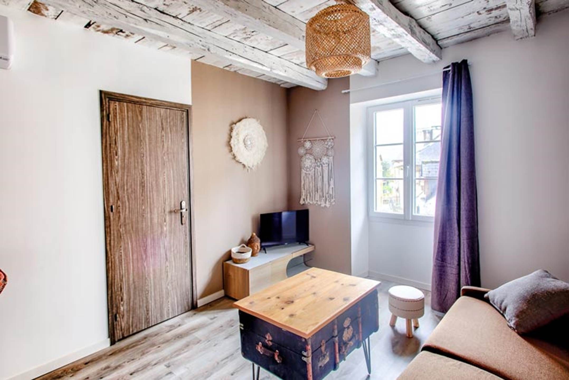 A room with a wooden door, a television on a stand, a small table, a sofa, and a window with curtains.