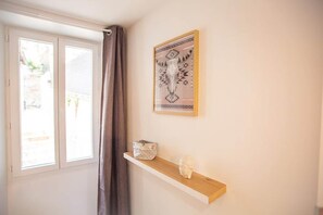 A room with a window, a curtain, a wall-mounted shelf with a decorative item, and a framed picture.
