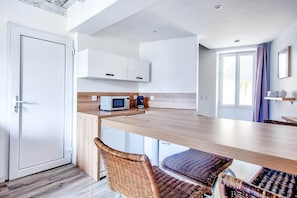 Eine moderne Küche mit weißen Schränken, einer Mikrowelle und einer Kaffeemaschine. Es gibt Korbstühle und einen Holztisch.