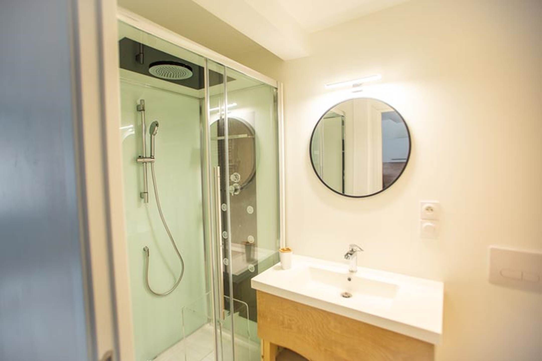 A bathroom with a glass-enclosed shower, a round mirror, and a white sink.