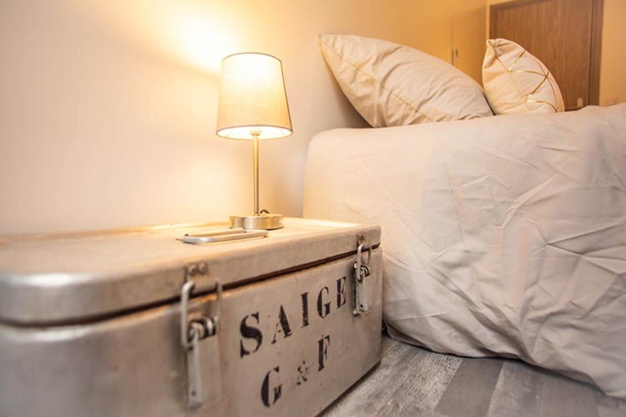 Un lit avec une couette blanche, une lampe de chevet et une valise sur laquelle on peut lire l’inscription « SAIGE G & F ».