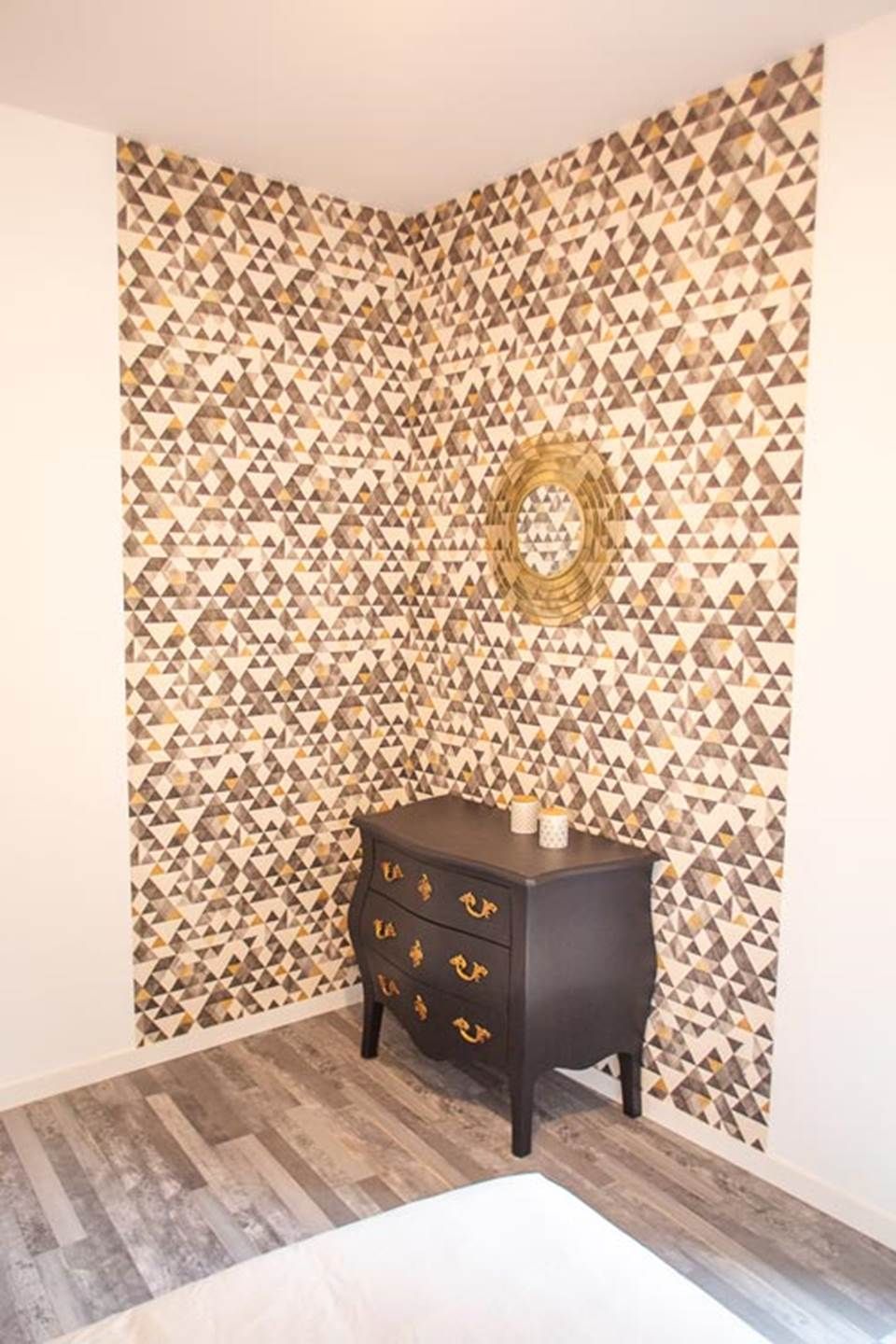 Una habitación de esquina con un papel tapiz estampado, una mesita de noche de madera oscura con cajones y suelo de madera.