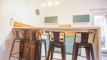Une cuisine avec un coin bar, quatre tabourets, des luminaires suspendus et une fenêtre.