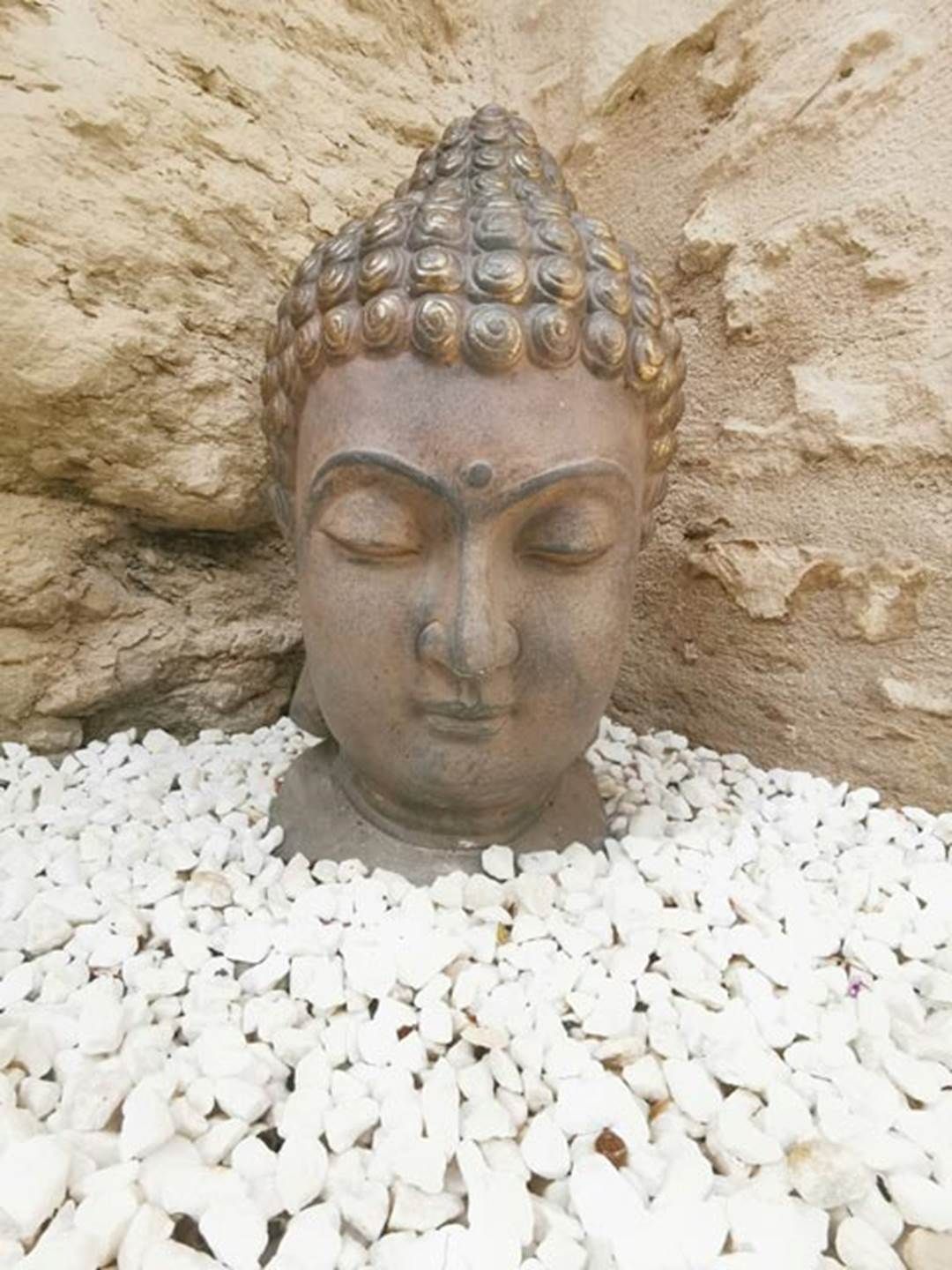 Une sculpture représentant la tête d’un Bouddha, en pierre, entourée de petits cailloux blancs.