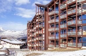 Un hôtel à plusieurs étages doté de balcons, une station de ski en arrière-plan et une enseigne indiquant « Supermarché Shepa ».