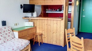 Une pièce compacte comprenant un lit superposé, une petite kitchenette, un bureau avec un ordinateur, une chaise et un lit avec une literie à motifs.