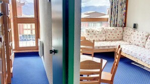 Une pièce avec un sol à pois bleus, une table en bois avec deux chaises, un canapé agrémenté de coussins à motifs et une vue sur les montagnes par la fenêtre.