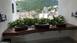 Une salle de bain dotée d’une grande fenêtre, de trois vasques en pierre et offrant une vue sur un paysage urbain avec des montagnes.