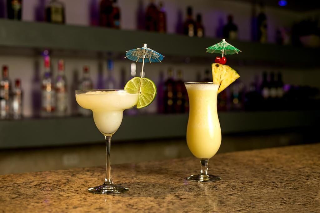 Two cocktails on a bar counter with a blurred background of shelves with bottles.
