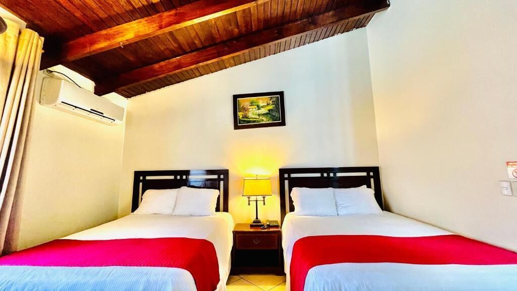 A room with two beds, a nightstand with a lamp, a painting on the wall, and an air conditioning unit.