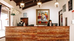 Un comptoir d’accueil avec deux employés, un grand tableau encadré au-dessus et un lustre.