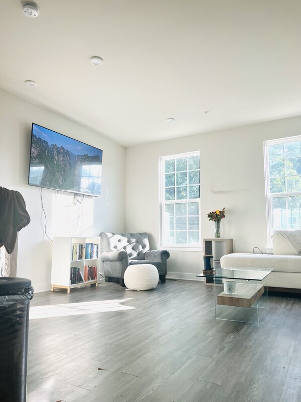 A modern living room with a flat-screen TV, a grey tufted armchair, a white sofa, and a glass coffee table.