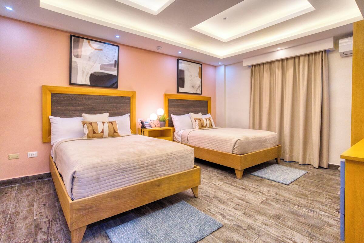 A hotel room with two beds, wooden headboards, a desk, and a large window with curtains.