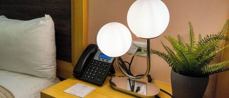 Una habitación de hotel con una mesita de noche que tiene un teléfono, una lámpara y una planta en maceta.