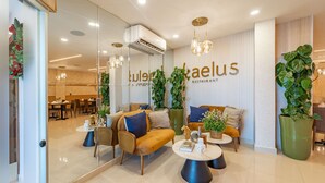 A modern restaurant interior with a seating area featuring sofas and round tables, a large potted plant, and a wall-mounted sign. - Melina Hotel (Higuey)