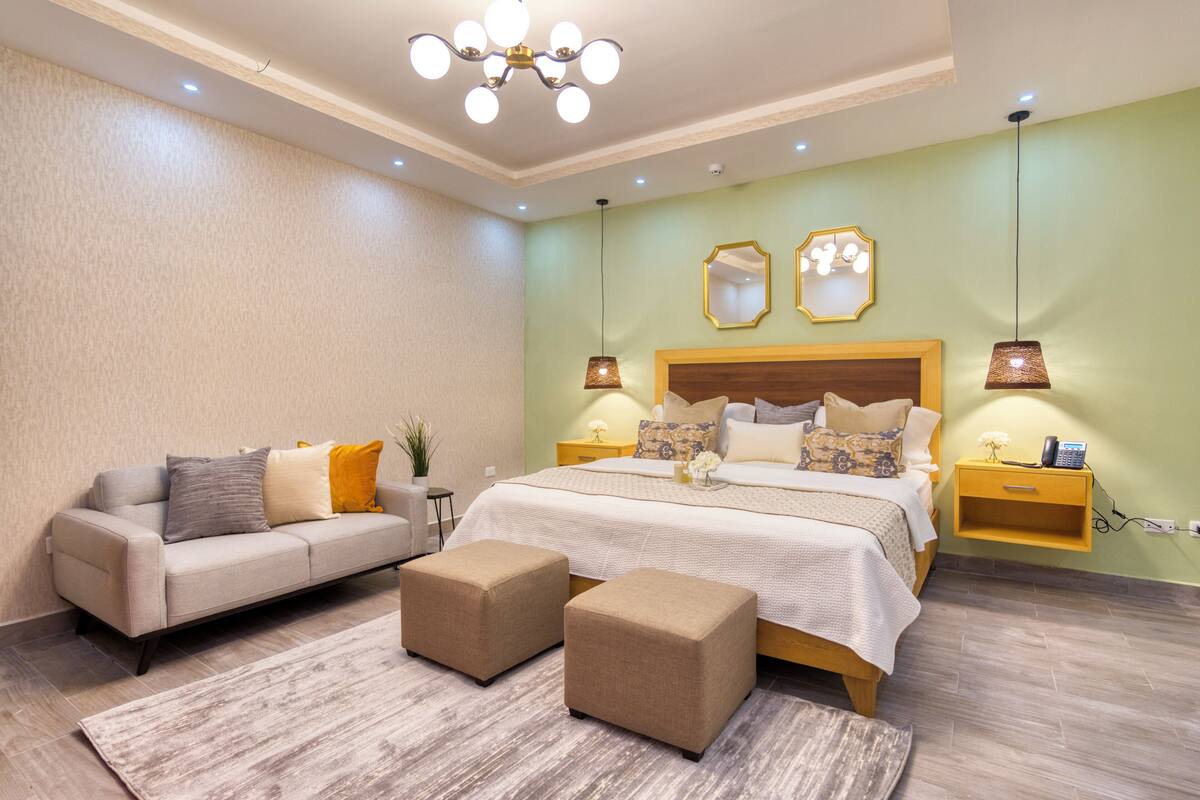 A modern bedroom with a large bed, a sofa, two ottomans, a nightstand, and a wall-mounted mirror.