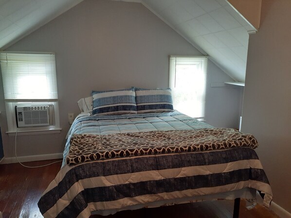 A bedroom with a sloped ceiling, a bed with a striped bedspread, two windows with blinds, and an air conditioning unit.