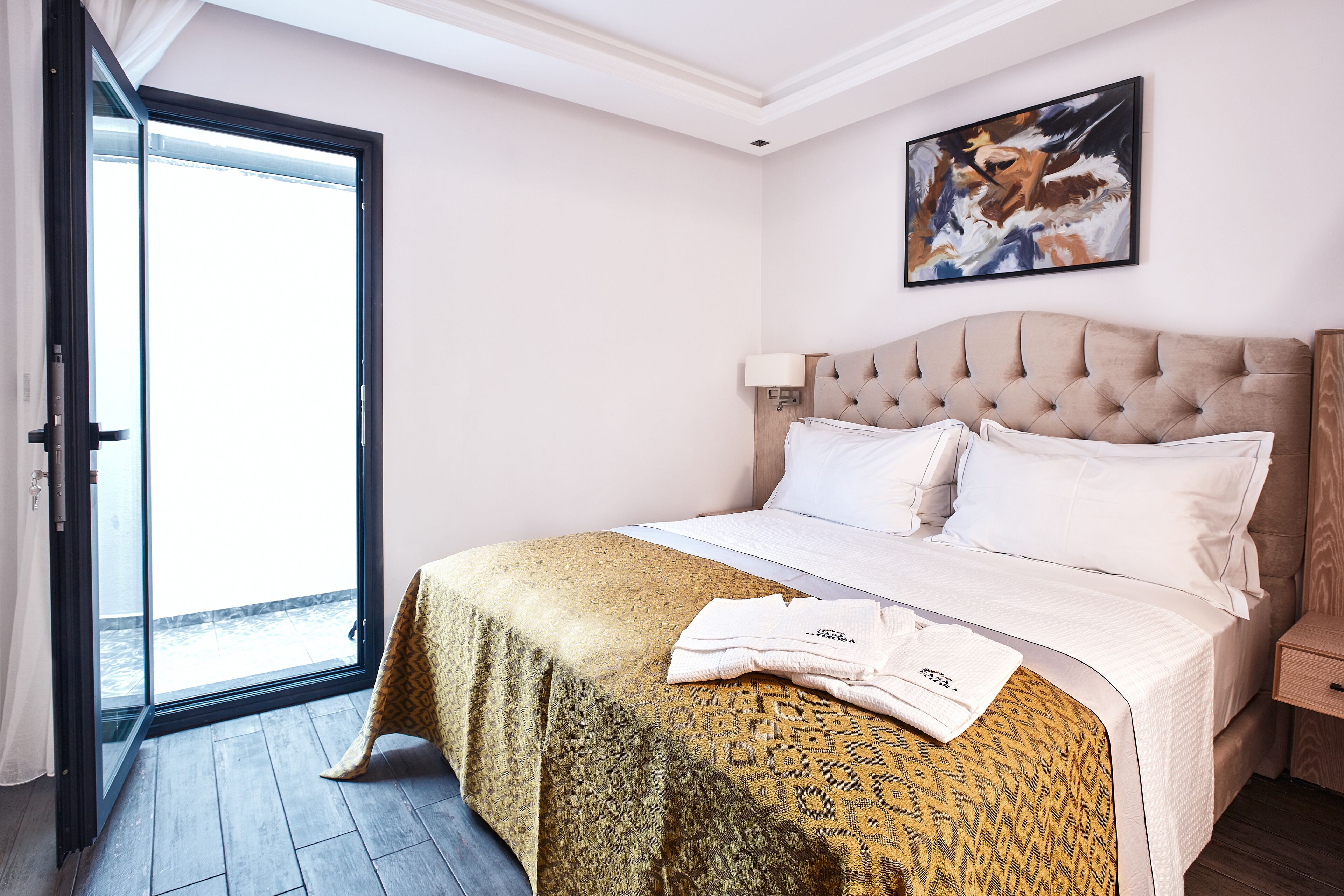 A hotel room with a bed, a nightstand, a window with a view, and a painting on the wall.