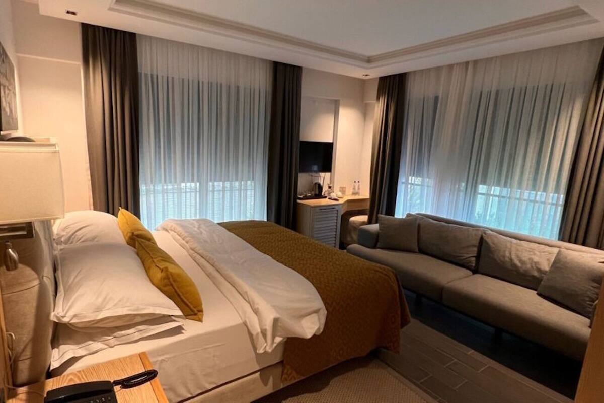 A hotel room with a bed, a sofa, a desk with a lamp, and a TV.