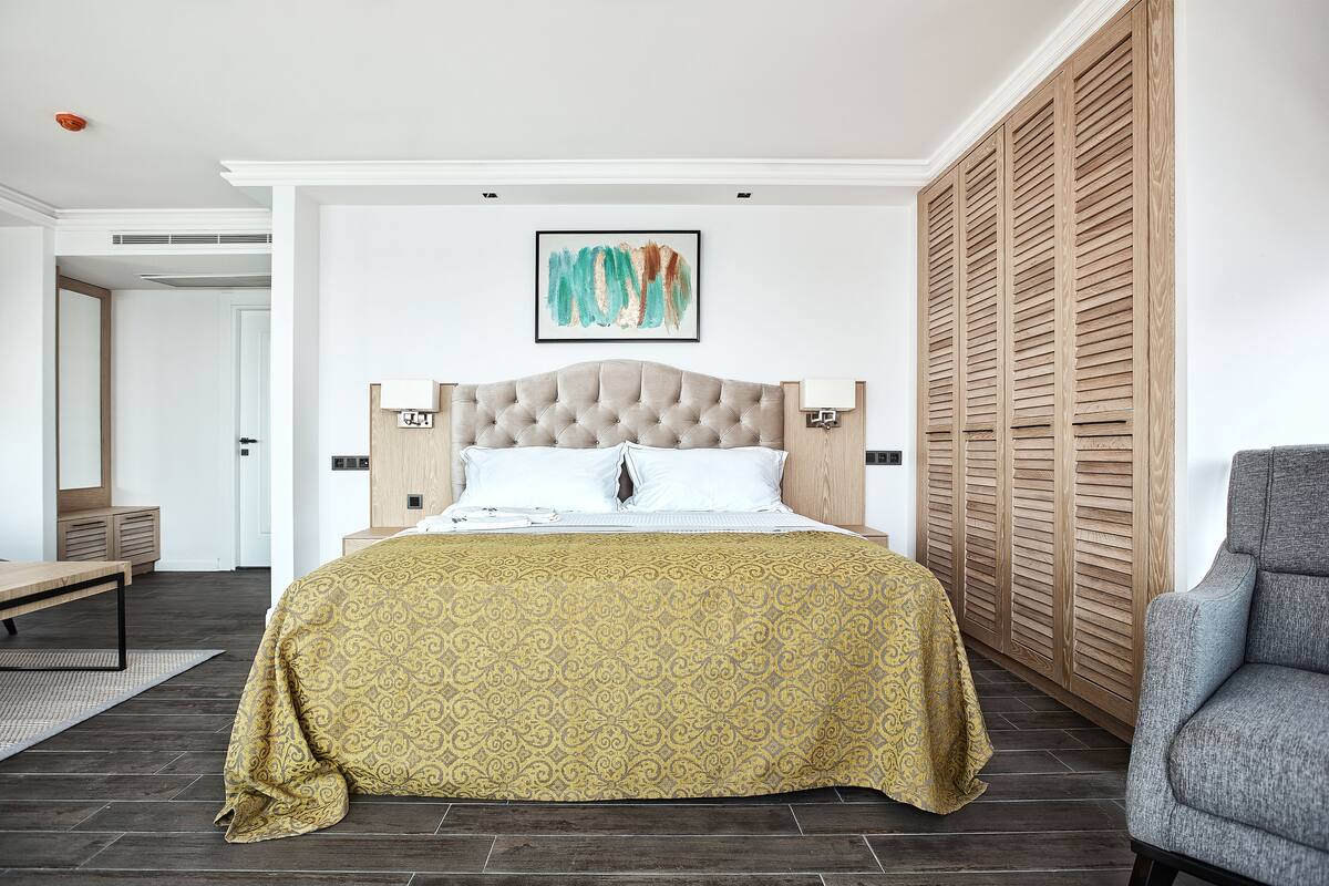A bedroom with a tufted headboard bed, a wooden wardrobe, and a grey armchair.