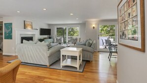 A modern living room with a fireplace, grey sofas, a white coffee table, and a dining area with outdoor seating. - Crystal Lake Waterfront Cottage (Orleans)
