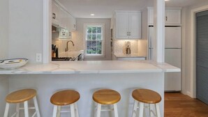 A modern kitchen with a central island, bar stools, and a view into the dining area. - Crystal Lake Waterfront Cottage (Orleans)