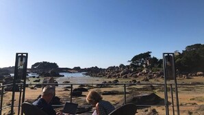 A beachside seating area with two people using laptops, overlooking a rocky shoreline with a clear sky. - APARTMENT "ILE BONO" WITH GARDEN, 800 ml BEACH (Perros-Guirec)