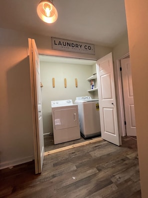 A laundry room with a sign reading "LAUNDRY CO.", a washing machine, a dryer, and wooden shelves with items on them. - Lakefront • Hot Tub • Private Dock • Kayaks • Canoe • Fire Pit • Dog Friendly (Coolbaugh Township)