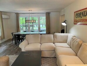 Una sala de estar con un sofá seccional color crema, una mesa de comedor con sillas, una araña y un letrero que dice 'Cornucopia Deli & Catering'.
