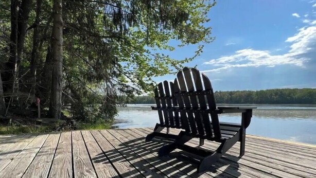 A wooden deck with a black Adirondack chair facing a calm lake, surrounded by trees. - Lakefront • Hot Tub • Private Dock • Kayaks • Canoe • Fire Pit • Dog Friendly (Coolbaugh Township)