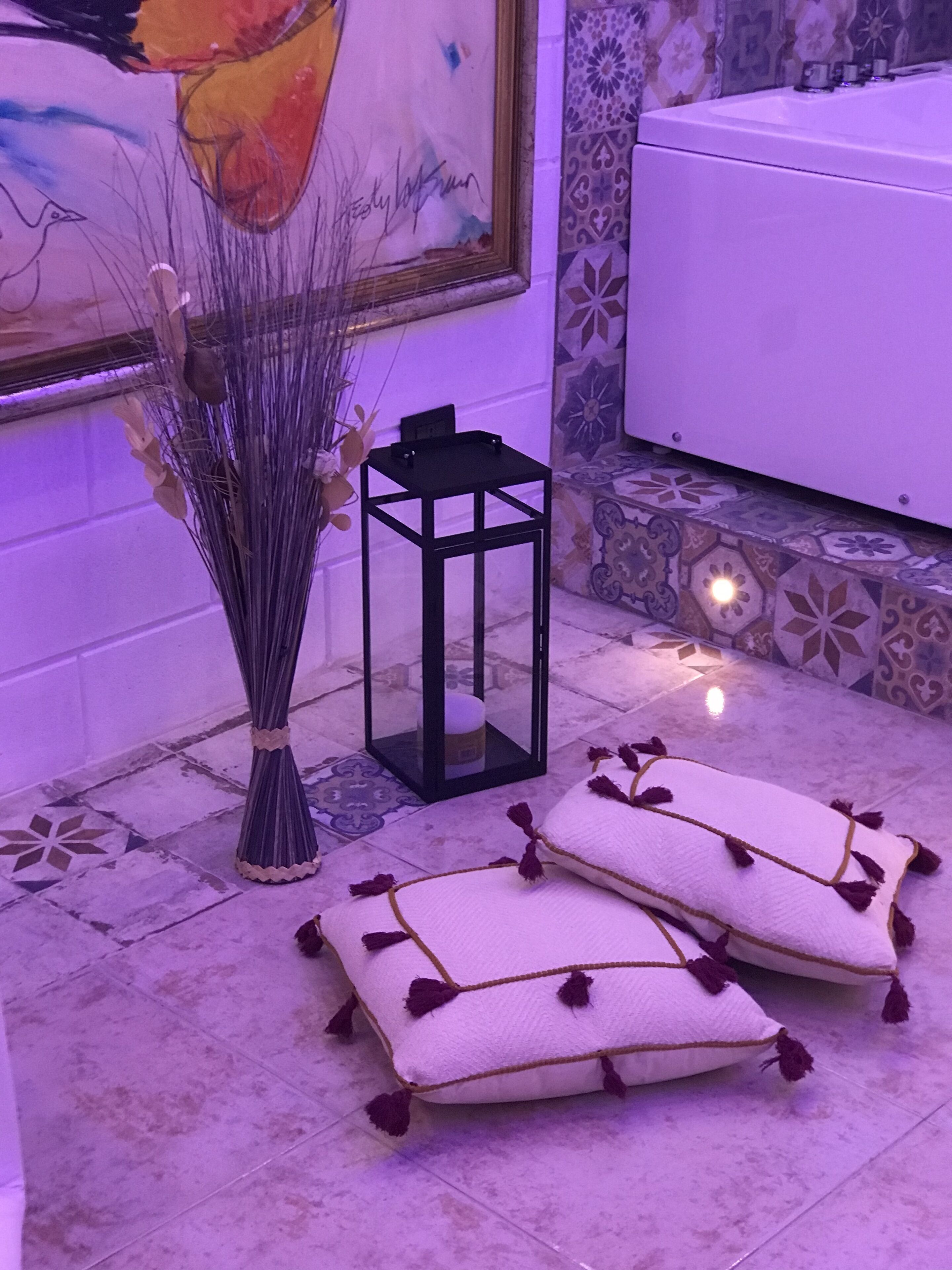 Une pièce avec un sol carrelé, une lanterne noire, un vase contenant des plantes séchées et deux coussins blancs ornés de glands.