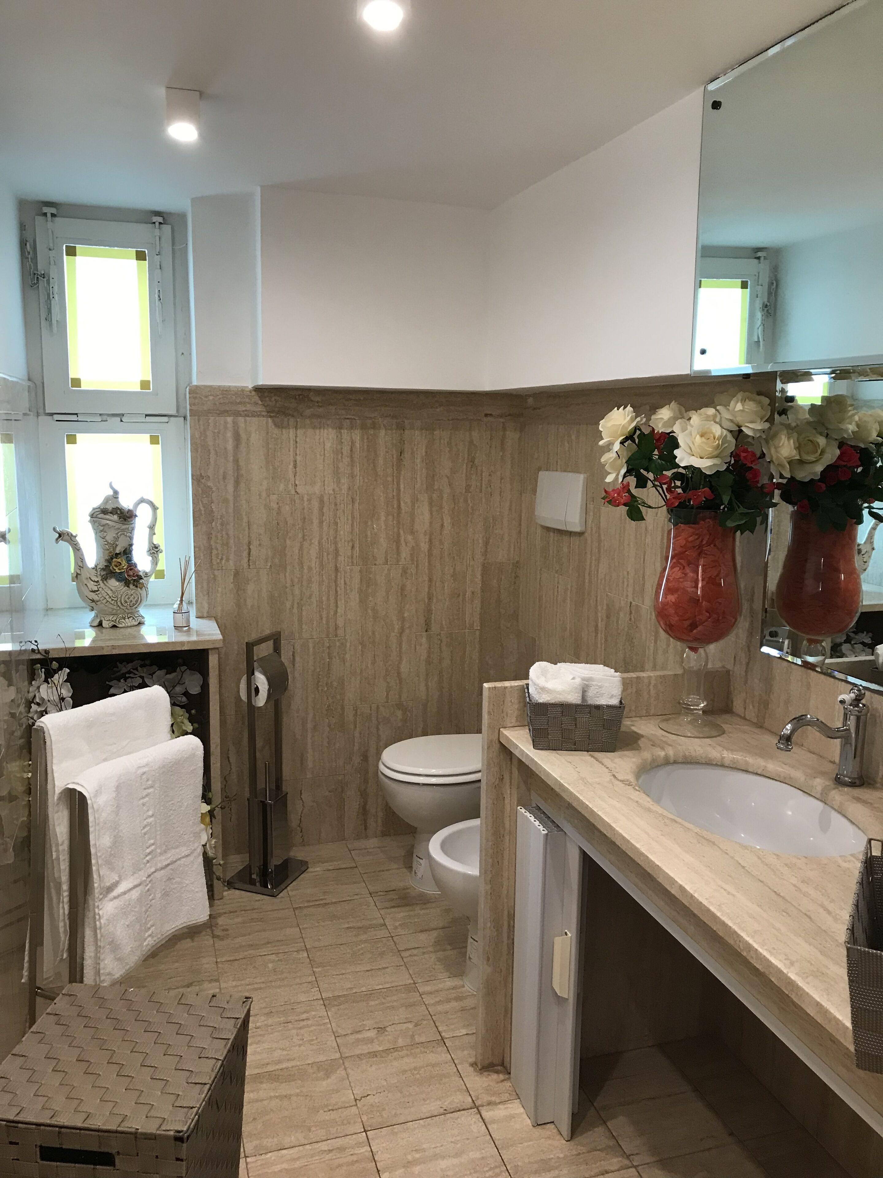 Un baño con piso de baldosas color beige, un lavamanos blanco, un espejo y dos floreros.