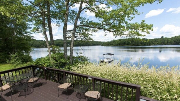 A deck with outdoor furniture overlooking a calm lake, surrounded by trees. - Charming Lake Front Cottage (Swan Lake)