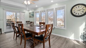 Une salle à manger avec une table et des chaises en bois, de grandes fenêtres, un ventilateur de plafond et une horloge murale.