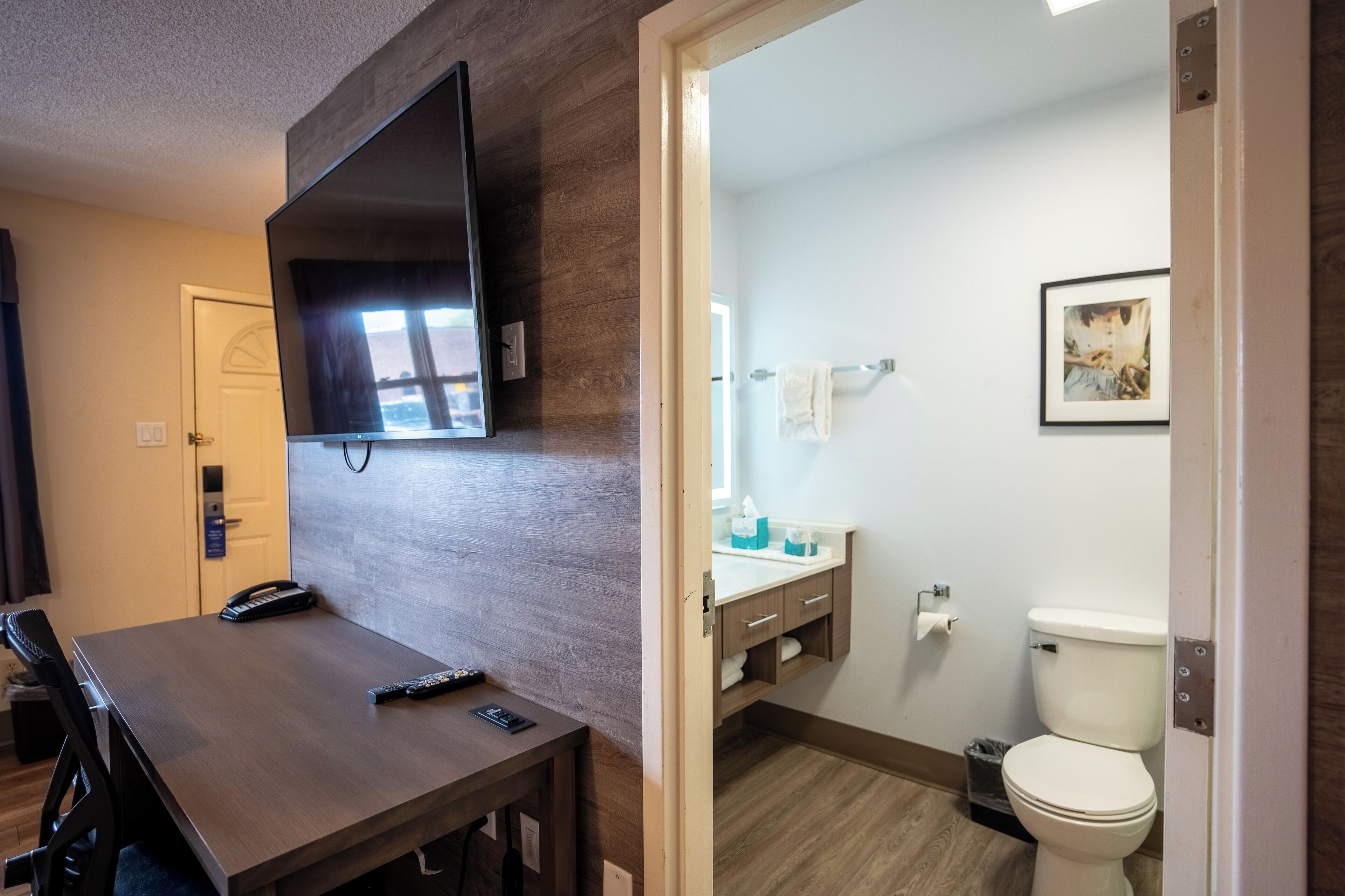 Habitación de hotel con una mesa de madera, un televisor de pantalla plana montado en la pared y un baño con inodoro, lavabo y toallero.