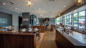 L’intérieur d’un restaurant est aménagé avec des tables et des chaises en bois, un coin bar et de grandes fenêtres donnant sur la rue.