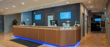 A reception desk with a wooden finish and blue LED lighting. There are two monitors on the wall displaying information.
