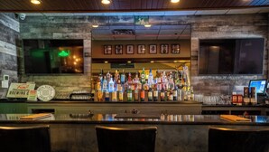 Un espace bar avec un comptoir où sont exposées différentes bouteilles et différents verres, des tableaux encadrés au mur et trois chaises au premier plan.