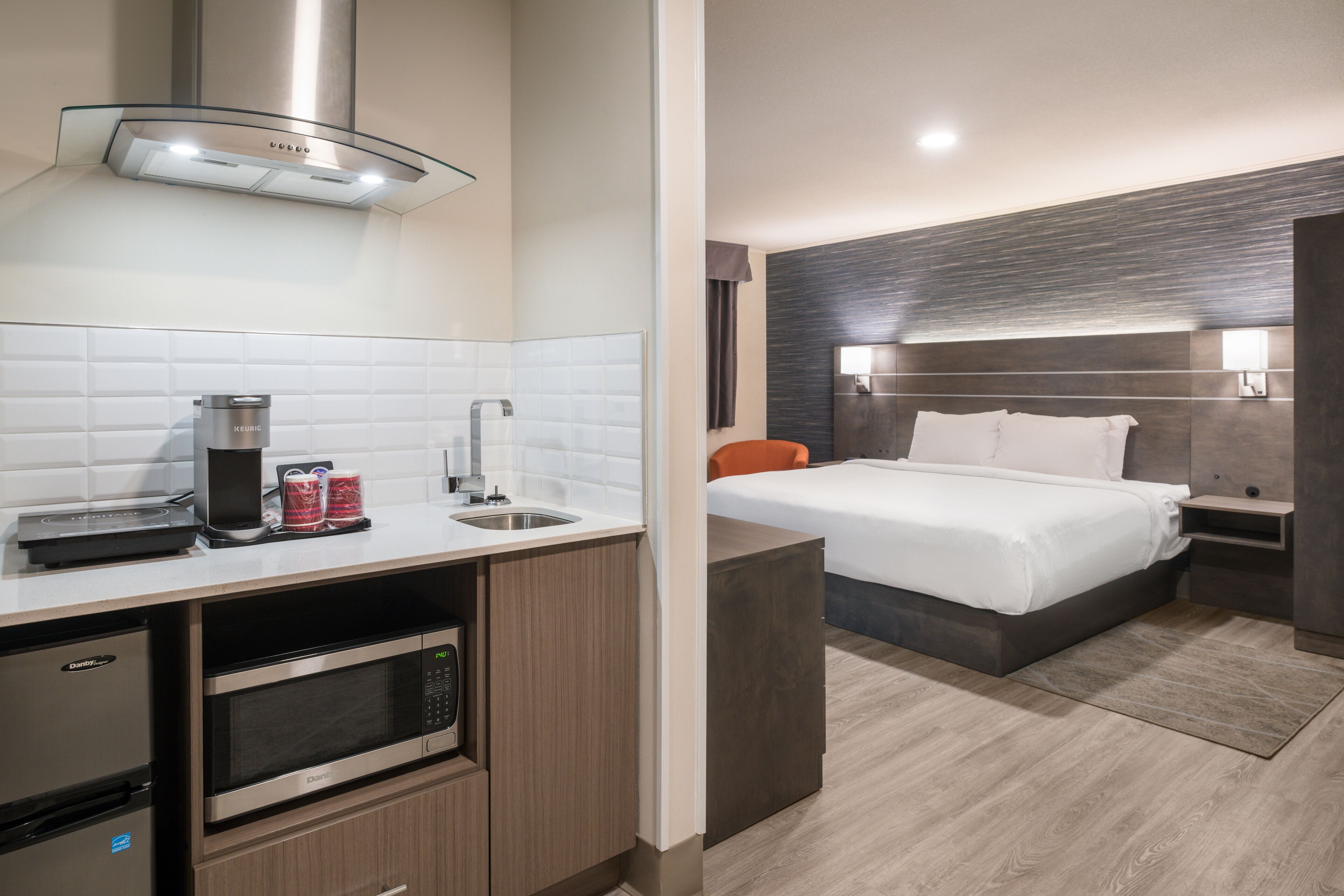 Una habitación de hotel moderna con kitchenette, una cama con ropa blanca, cabecera de madera oscura y una mesita de noche pequeña.