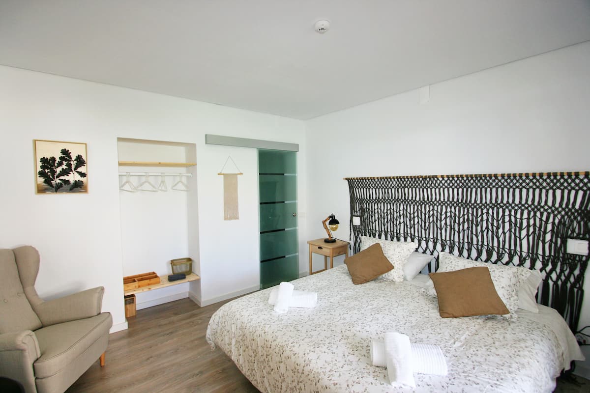 Un dormitorio con cama, una silla, una estantería con ropa colgada y una puerta que da a otra habitación.