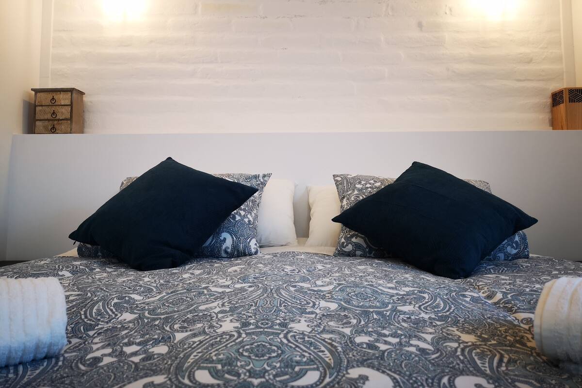 Una cama con una colcha estampada, dos almohadas negras y una mesita de noche de madera con un cajón.