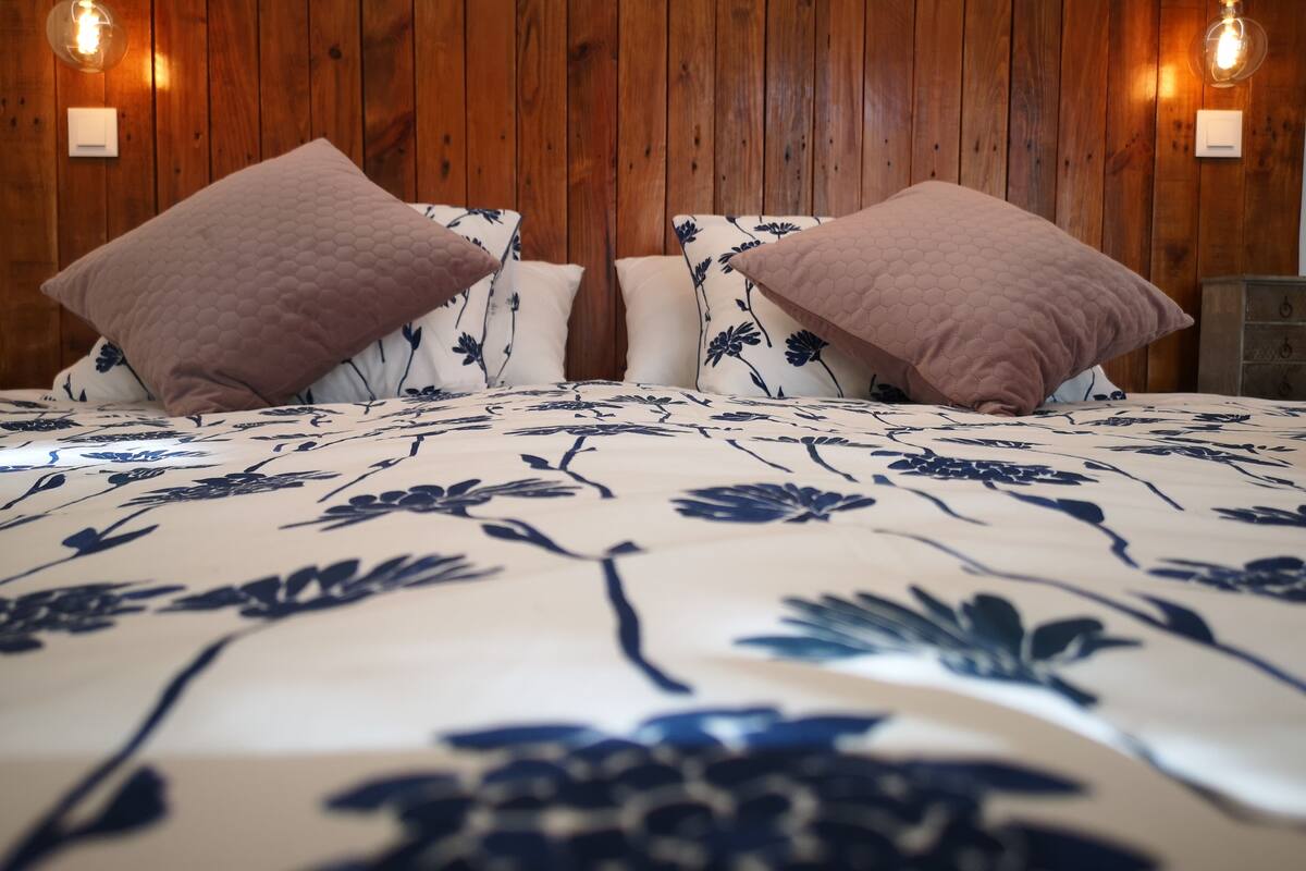 Una cama con ropa de cama floral, paredes de paneles de madera y dos lámparas fijadas a la pared.