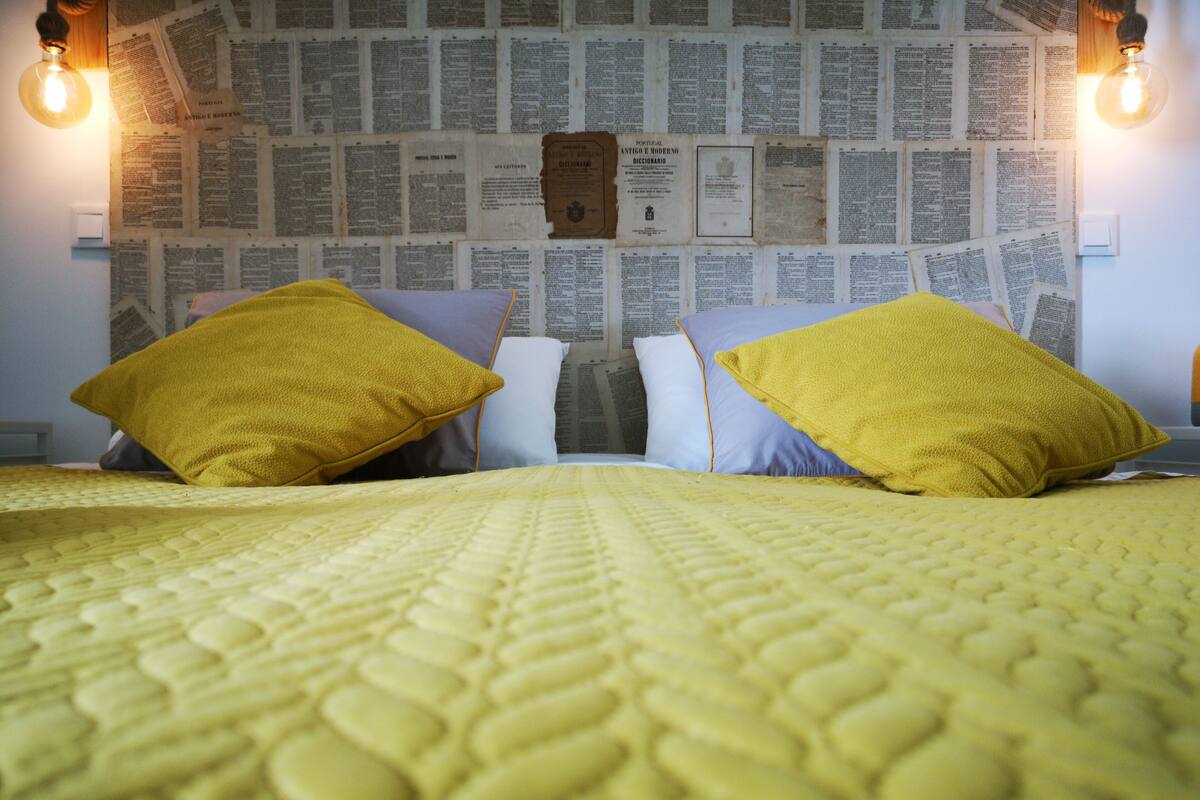 Una cama con un edredón amarillo, dos almohadas y una pared cubierta de recortes de periódico.