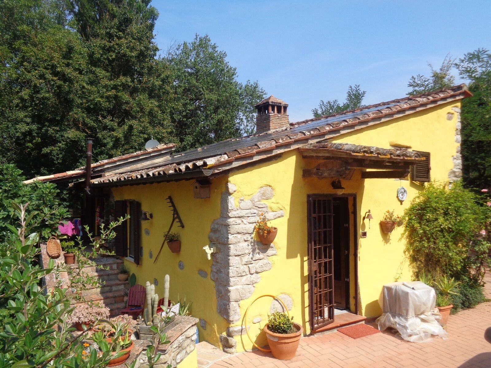 Un petit bâtiment jaune avec un mur en pierre, un toit en tuiles et une porte. On y trouve des plantes en pot et une petite table à l’extérieur.