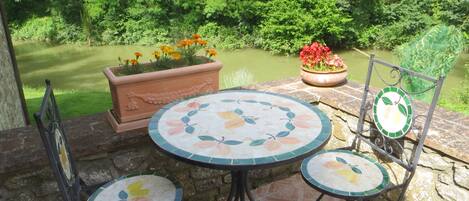 Un patio con una mesa redonda y dos sillas, rodeado de plantas en macetas y vistas a un estanque.